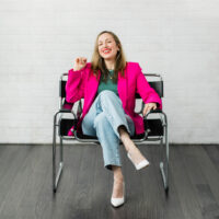 Woman sitting in a chair wearing a hot pink blazer green t shirt jeans and white heels.