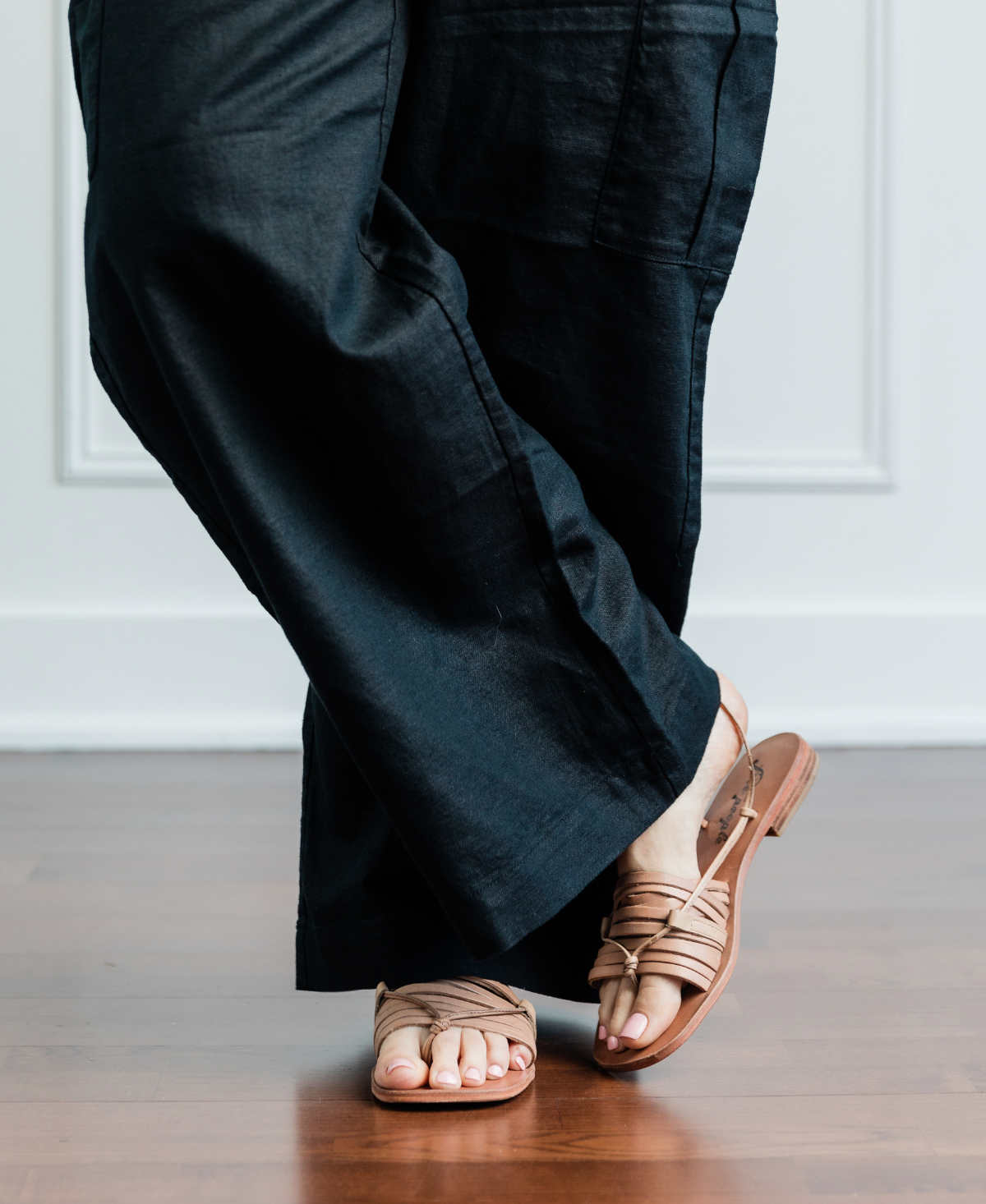 Cropped view of woman's legs wearing beige strappy sandals with full length wide leg black linen pants.