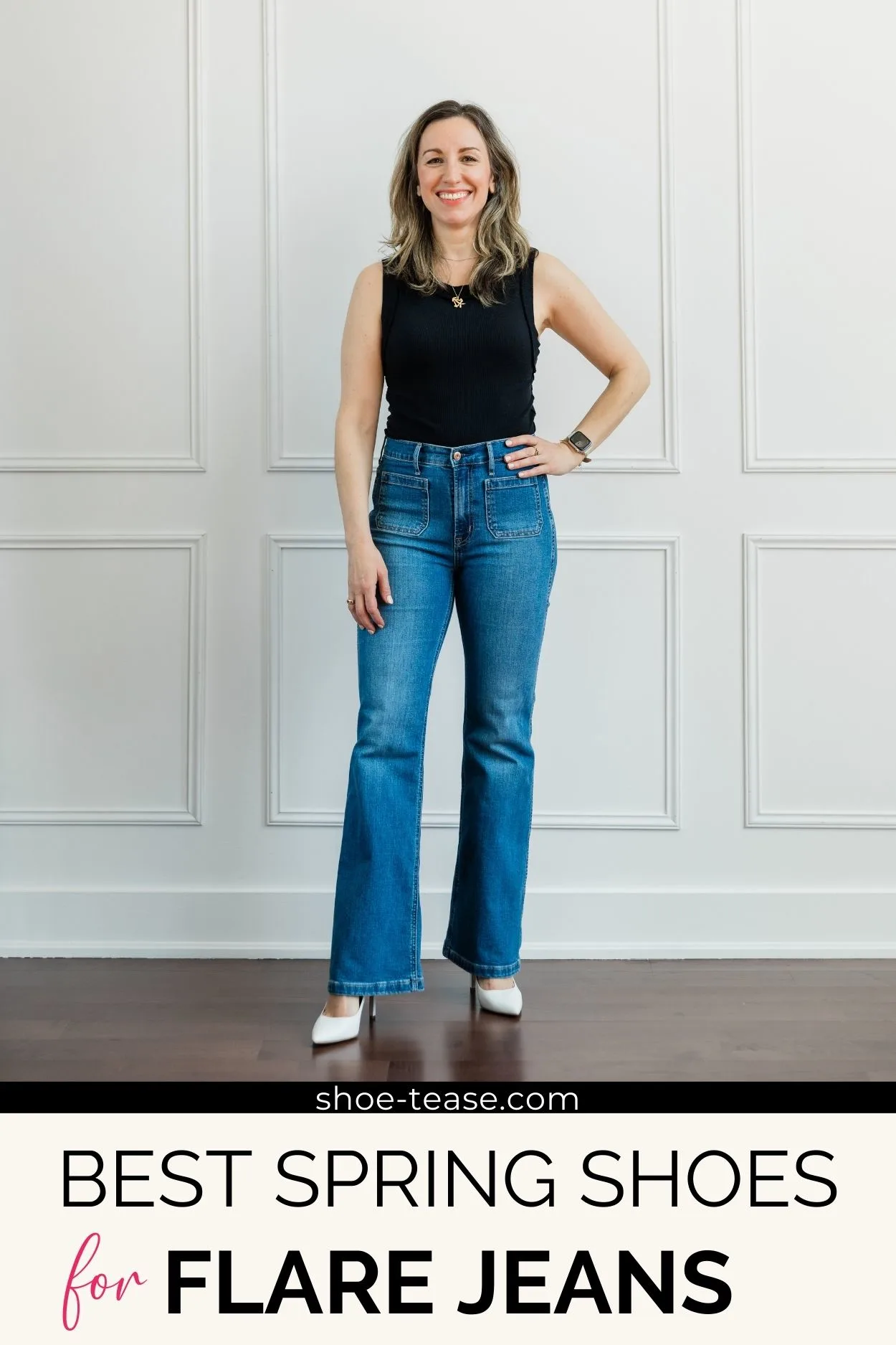 Woman posing wearing a black tank top, long blue denim flare jeans and white high heel pumps standing in front of a white wall.
