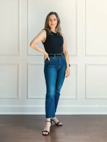 Woman posing in a black tank top, blue denim kick flares with black belt and sandals standing in front of a white wall.
