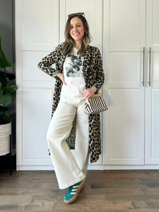 Brunette woman posing wearing off white wide leg jeans, graphic tee, green sneakers and leopard print trench coat.