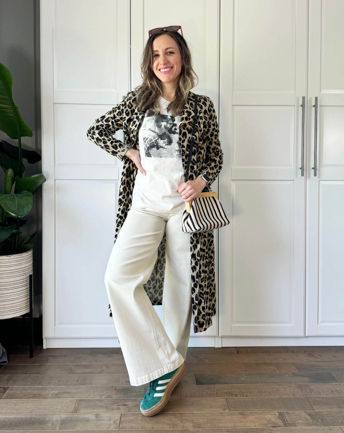 Brunette woman posing wearing off white wide leg jeans, graphic tee, green sneakers and leopard print trench coat.